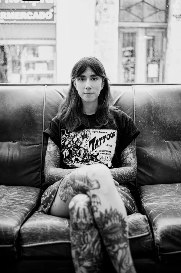 María sentada en el sofá de cuero del estudio, con las piernas tatuadas cruzadas.