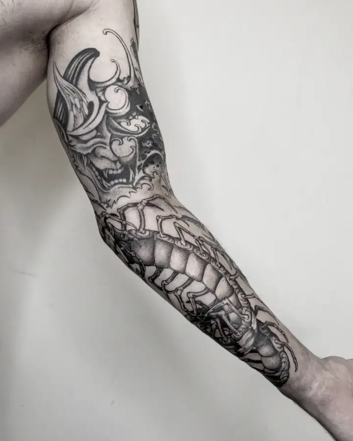 En el brazo estirado, una manga completa: máscara de demonio arriba y un ciempiés enorme por el antebrazo, en dotwork.