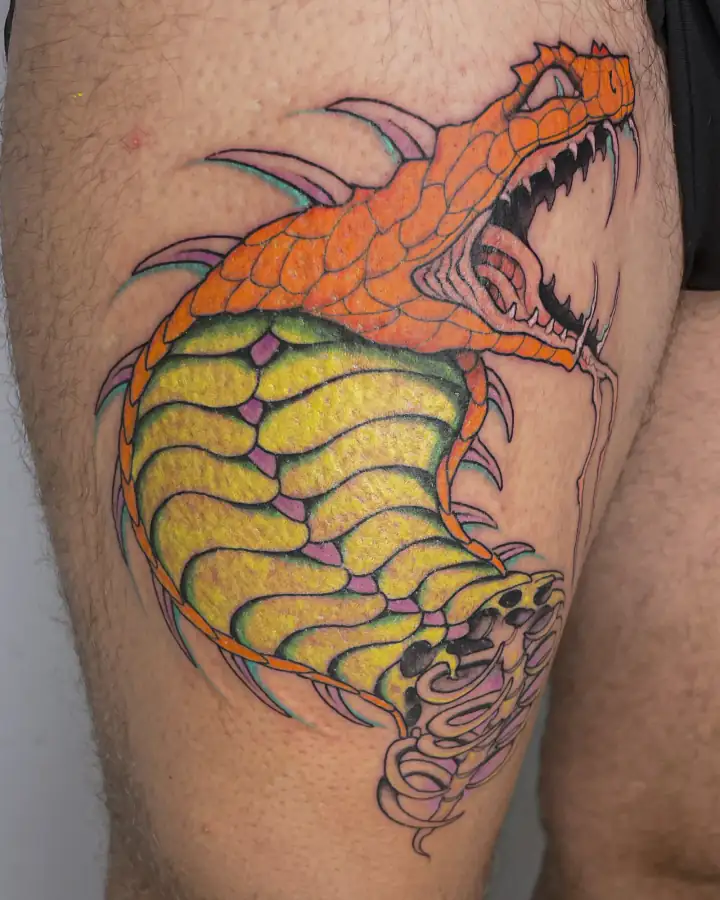 Muslo tatuado a color con una criatura escamosa que abre las fauces, en naranjas, amarillos y verdes.