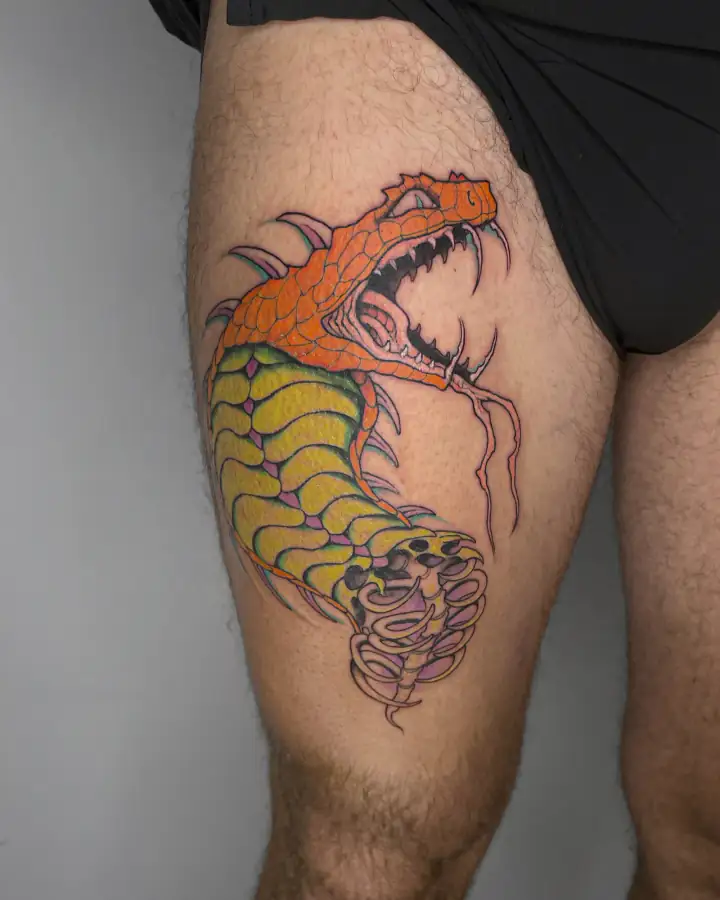 En el muslo, una criatura escamada naranja abre las fauces sobre un cuerpo amarillo; tatuaje a color.