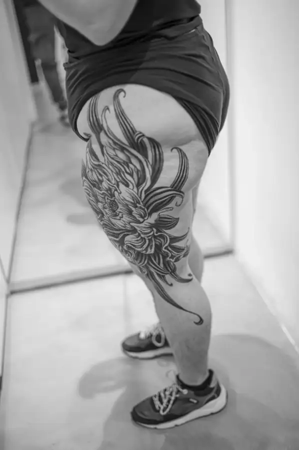 Perfil de la pierna con la flor que arranca en la cadera y se deshilacha en volutas sobre el muslo, en black & grey.