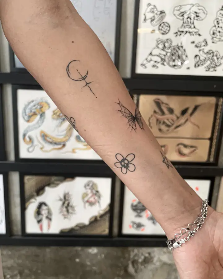 Antebrazo apoyado ante una pared de diseños, con una luna, una mariposa de telaraña y una flor a línea fina.