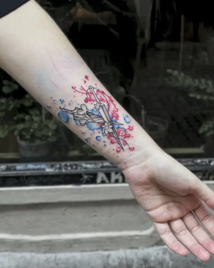 Antebrazo con una daga y flores dibujadas a línea fina sobre salpicaduras de acuarela azules y rojas.
