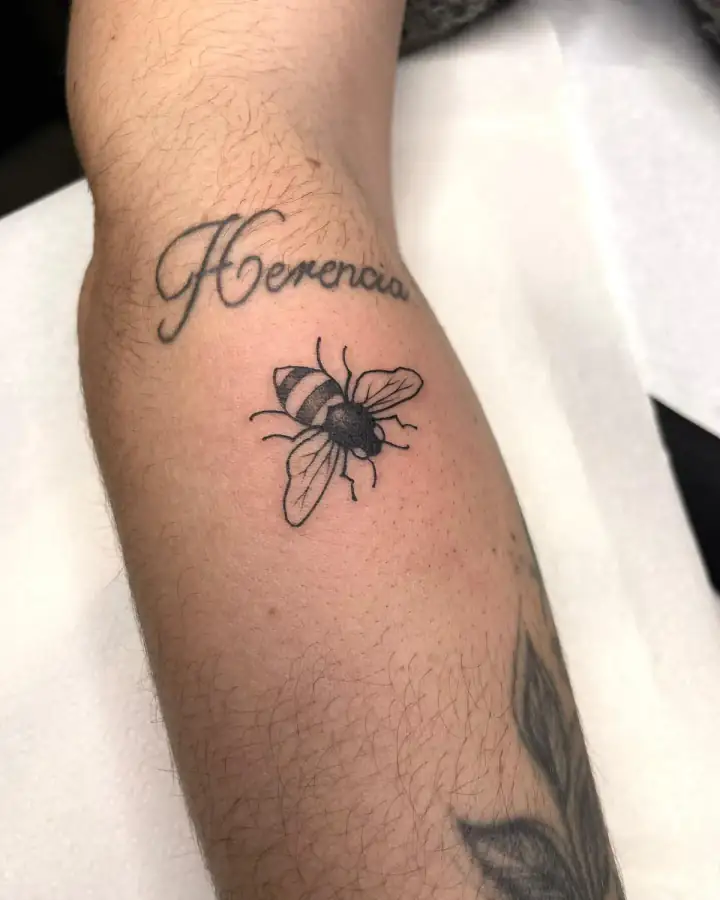 Antebrazo: una abeja de alas transparentes bajo la palabra «Herencia» en lettering, con línea fina y sombras suaves.