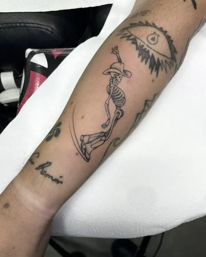 En el antebrazo, un esqueleto con sombrero y un brazo en alto se desliza sobre una línea curva, a línea fina y sombra suave.