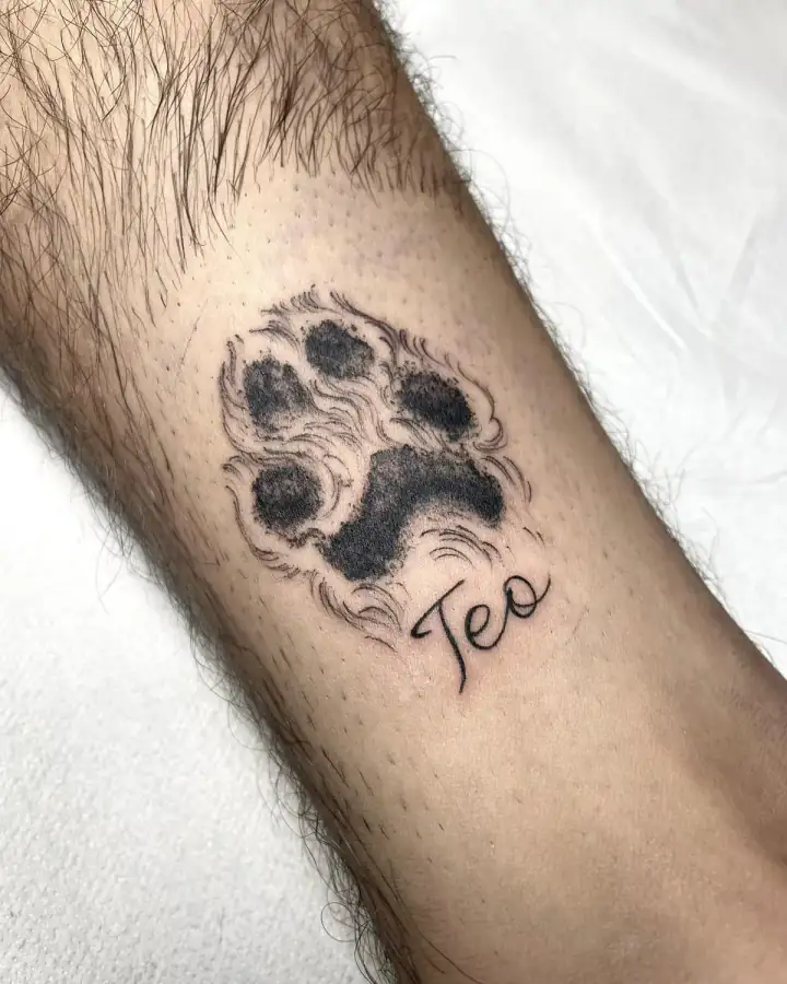 Antebrazo con la huella de un perro rellena en negro, sombreada a puntitos, y un nombre corto en lettering manuscrito.