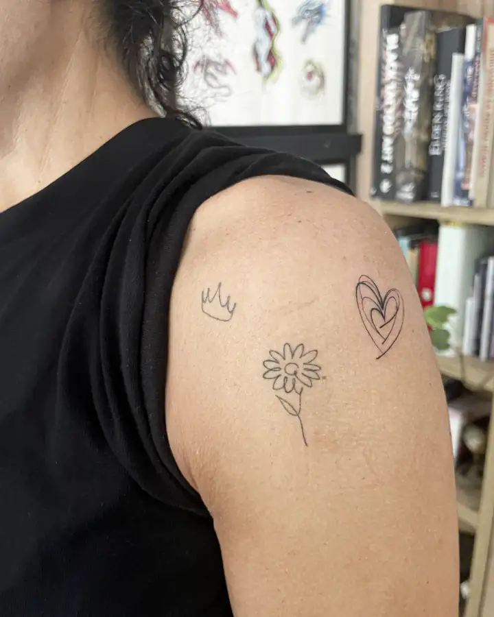 Hombro con tres dibujos pequeños de línea fina y trazo suelto: una corona, una margarita y un corazón.
