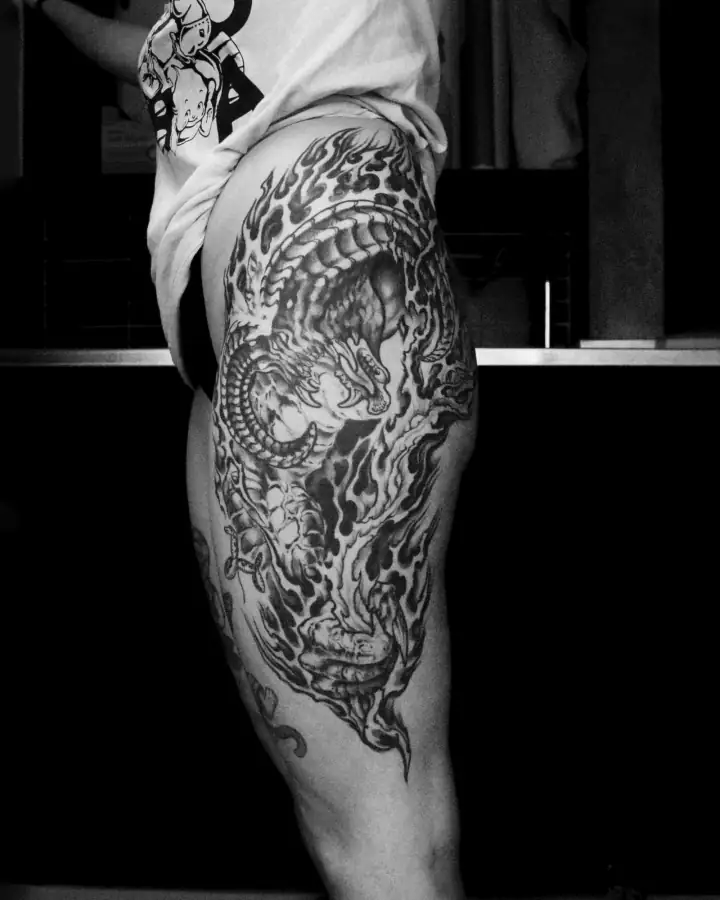 Cadera y muslo con un dragón enroscado entre llamas, en black & grey de fuerte contraste.