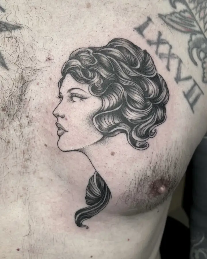 En el pecho, el perfil de una mujer de pelo ondulado en black & grey de sombras punteadas, junto a unas cifras romanas.
