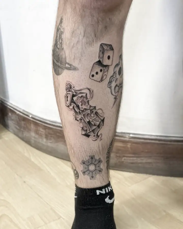 En la espinilla, varios tatuajes pequeños sueltos —dados, un planeta anillado y un adorno barroco— en black & grey.