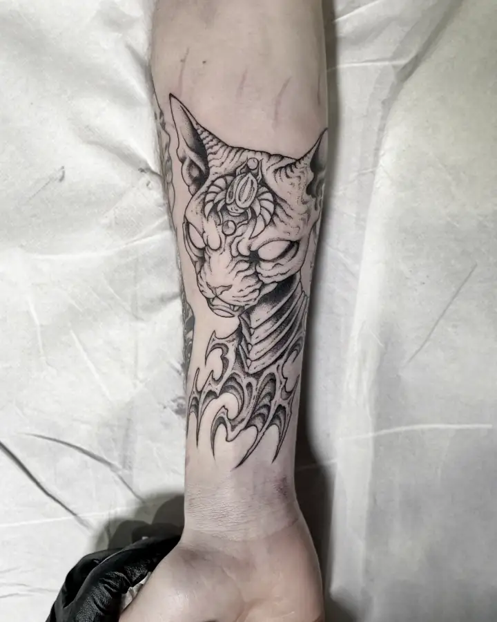 Antebrazo con la cabeza de un gato esfinge y una joya en la frente, sobre llamas neotribales en black & grey.