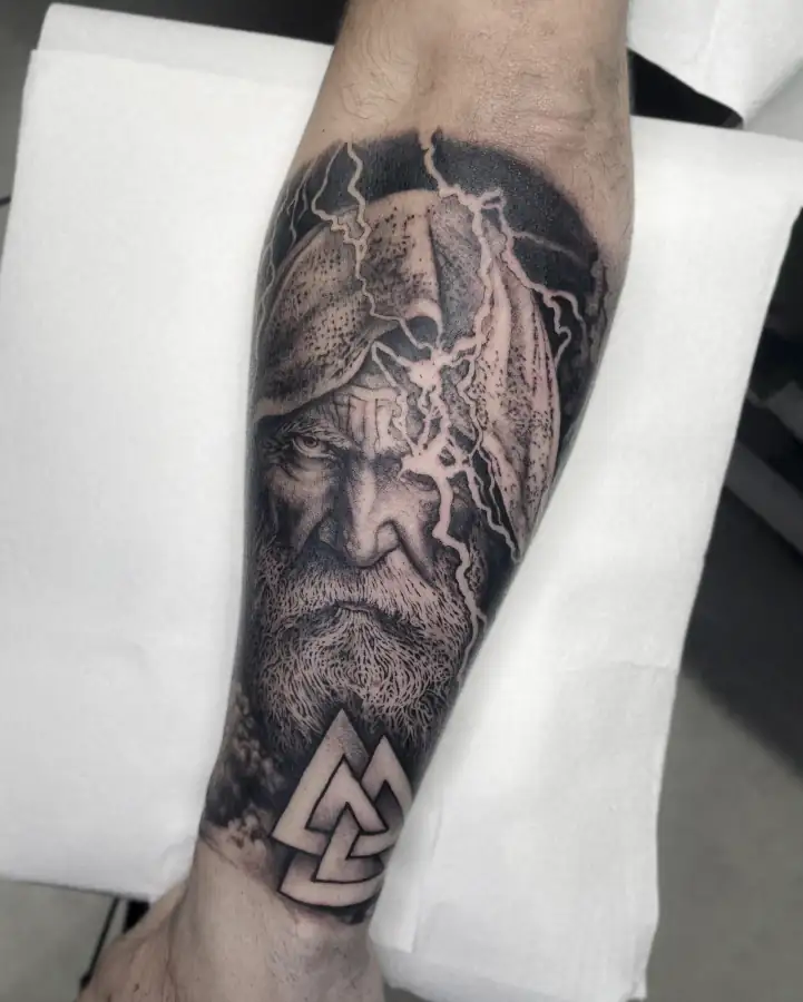 Pierna cubierta por el rostro de un anciano barbado encapuchado, rayos blancos y un valknut; realismo en black & grey.