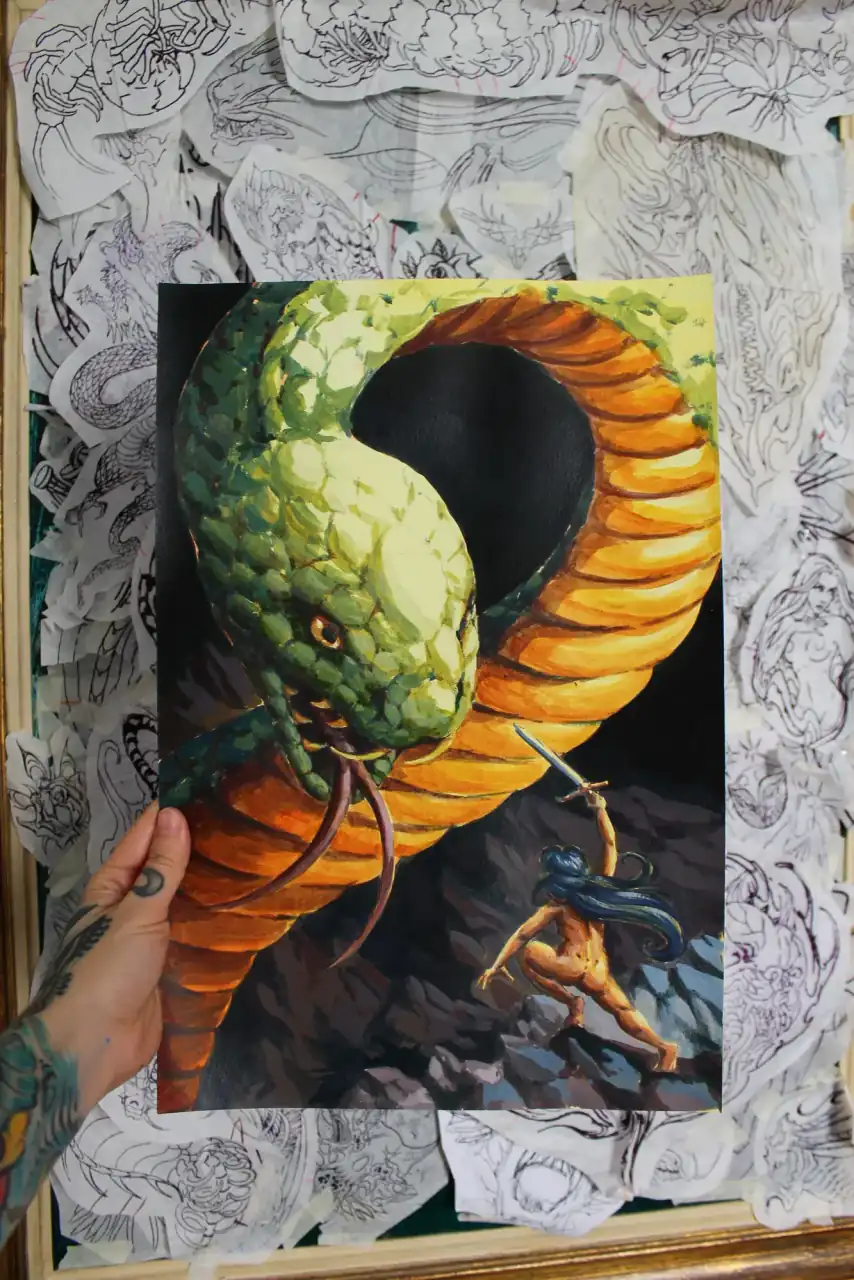 Lámina a color de InkSnake sostenida con una mano tatuada: una serpiente verde y naranja enroscada frente a una guerrera con espada, delante de una pared de dibujos.