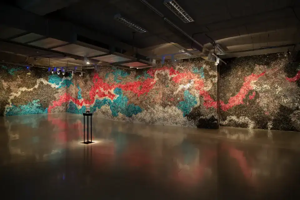 MUR MUR, pintura sobre papel (2018). Sala de exposiciones en penumbra con dos paredes cubiertas de pintura sobre papel en rojo, turquesa y pardo, y una peana iluminada en el centro que se refleja en el suelo pulido.
