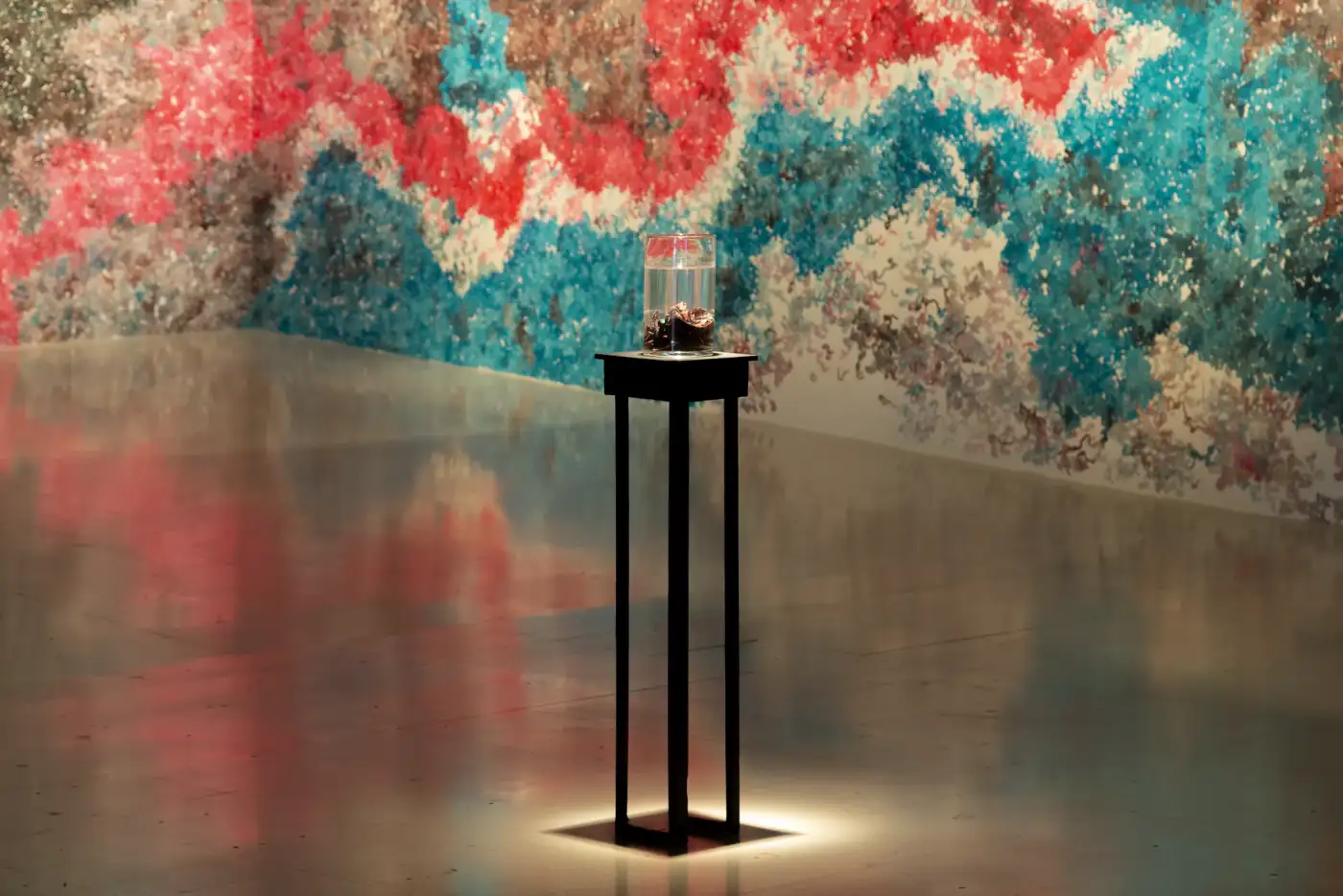 MUR MUR (2018). Peana negra alta con un cilindro de vidrio, iluminada desde arriba, delante del mural rojo y turquesa que se refleja en el suelo.