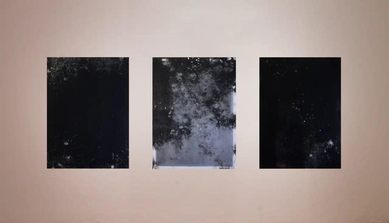 Tres obras verticales colgadas en fila sobre la pared de una sala: las laterales casi negras con leves texturas claras; la central, con zonas gris azuladas y manchas claras en la mitad inferior.
