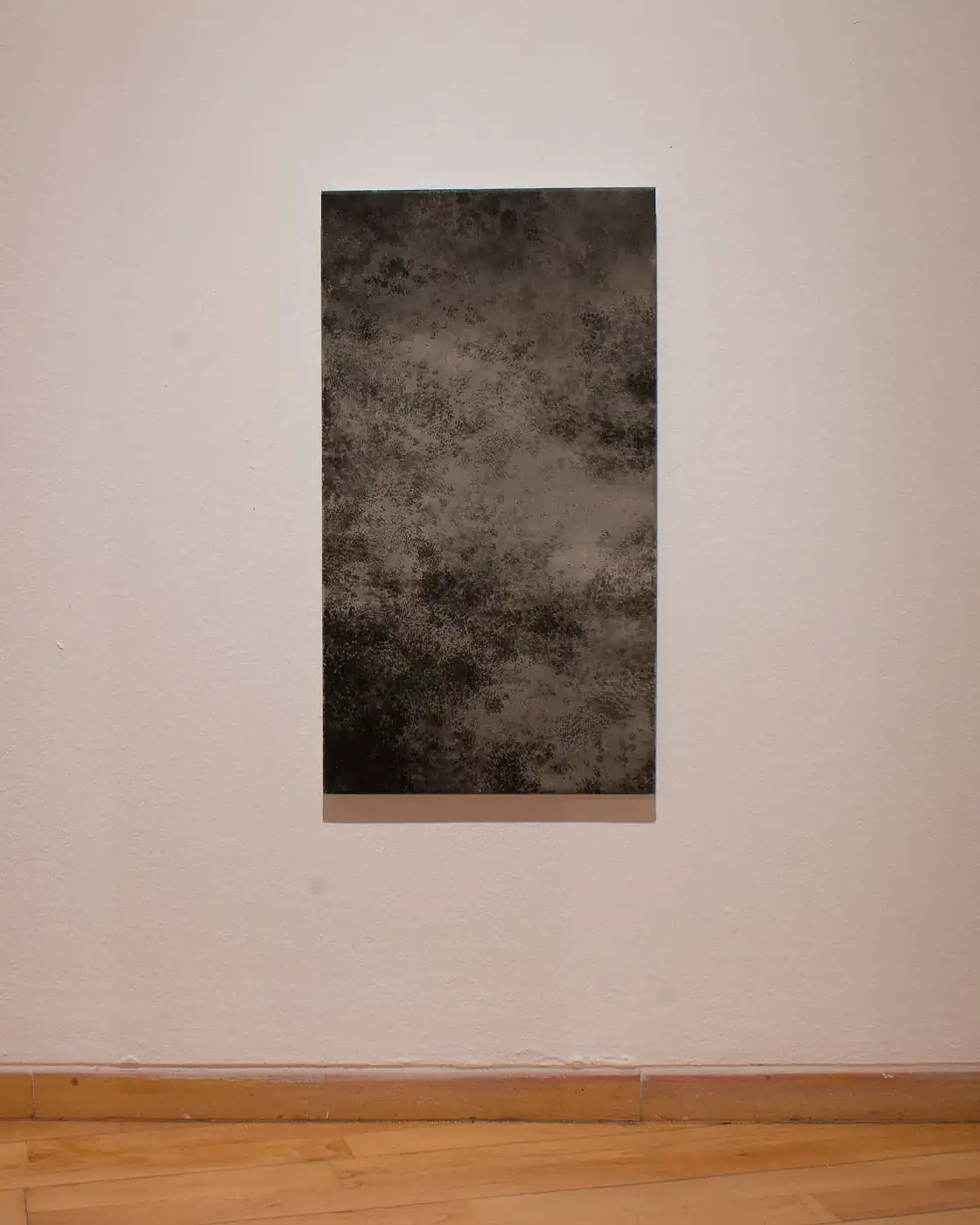 Pieza vertical de textura moteada gris y negra montada en la pared blanca de una sala, con zócalo y suelo de madera debajo.