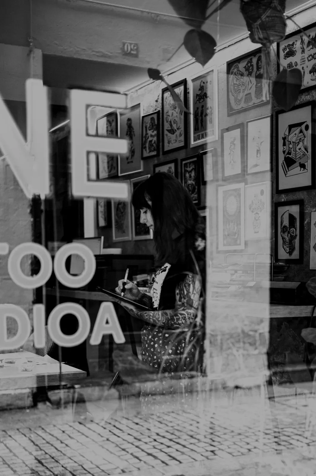 María dibujando con el lápiz sobre una tableta dentro del estudio, vista desde la calle a través del escaparate, con las letras del rótulo reflejadas en el cristal y las láminas de flash enmarcadas en la pared. Fotografía en blanco y negro.