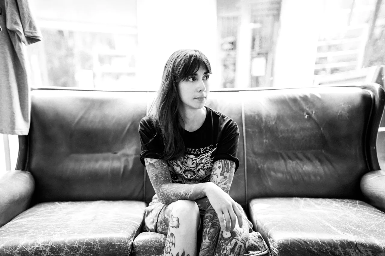 María sentada en el sofá de cuero del estudio, con la camiseta de InkSnake y la luz de la ventana detrás. Fotografía en blanco y negro.
