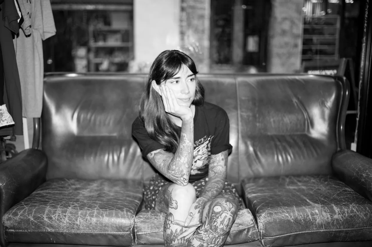 María sentada en el sofá de cuero marrón del estudio, con la mano en la barbilla y los brazos tatuados a la vista. Fotografía en blanco y negro.