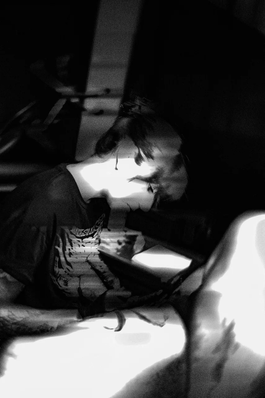María inclinada sobre la pierna de una clienta, tatuando muy cerca de la piel con la luz dura de la ventana. Fotografía en blanco y negro.