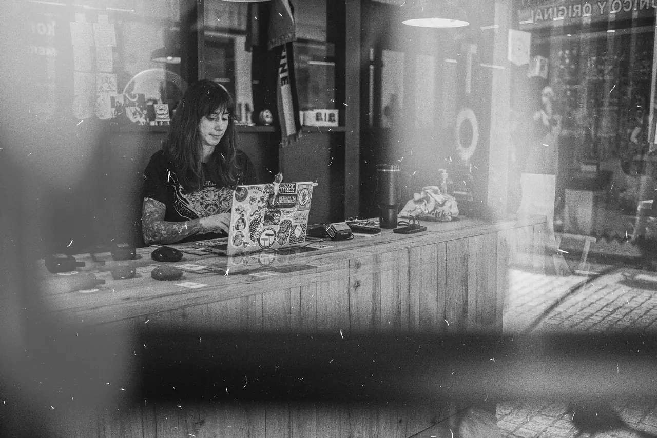 El mostrador de madera del estudio visto desde la calle a través del cristal, con María al portátil. Fotografía en blanco y negro.