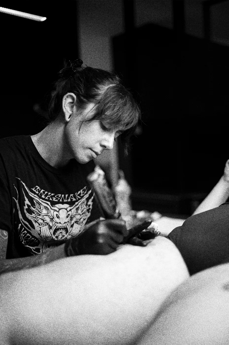 María tatuando la cadera de una clienta, con la camiseta de InkSnake. Fotografía en blanco y negro.