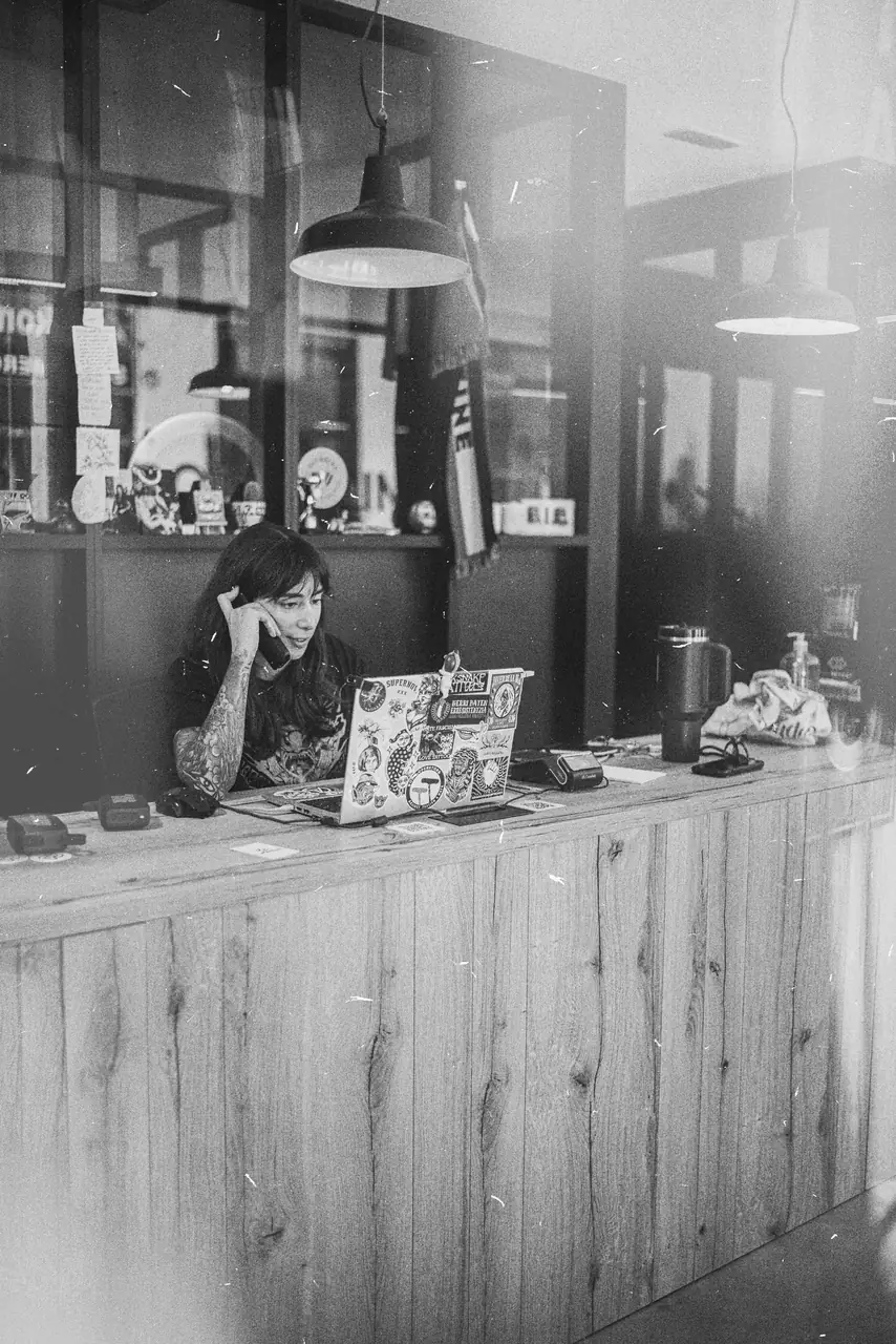 El interior del estudio desde la puerta, con María al portátil tras el mostrador de madera y los reflejos de la calle en el cristal. Fotografía en blanco y negro.