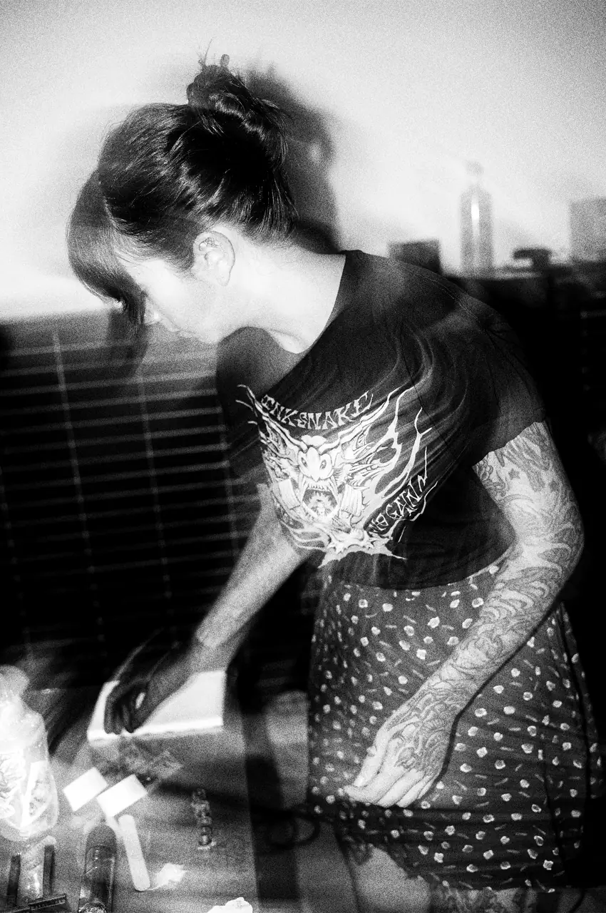 María Vera inclinada sobre la mesa de trabajo, con el pelo recogido, camiseta de InkSnake y los brazos tatuados, en una fotografía en blanco y negro movida y con doble exposición.