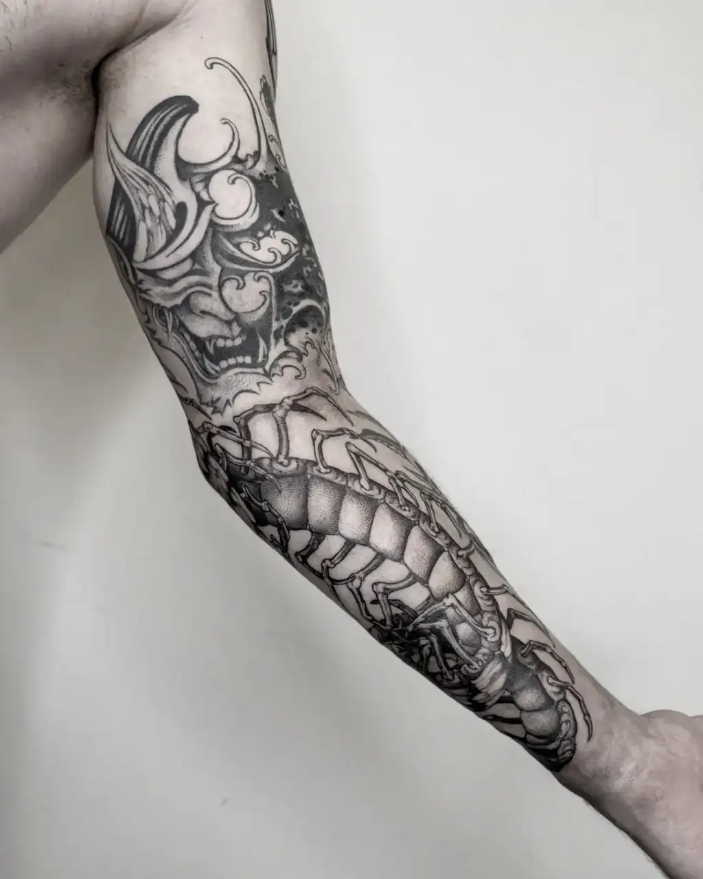 Brazo estirado con la manga entera: una máscara de demonio y un ciempiés gigante en black & grey sombreado con dotwork.