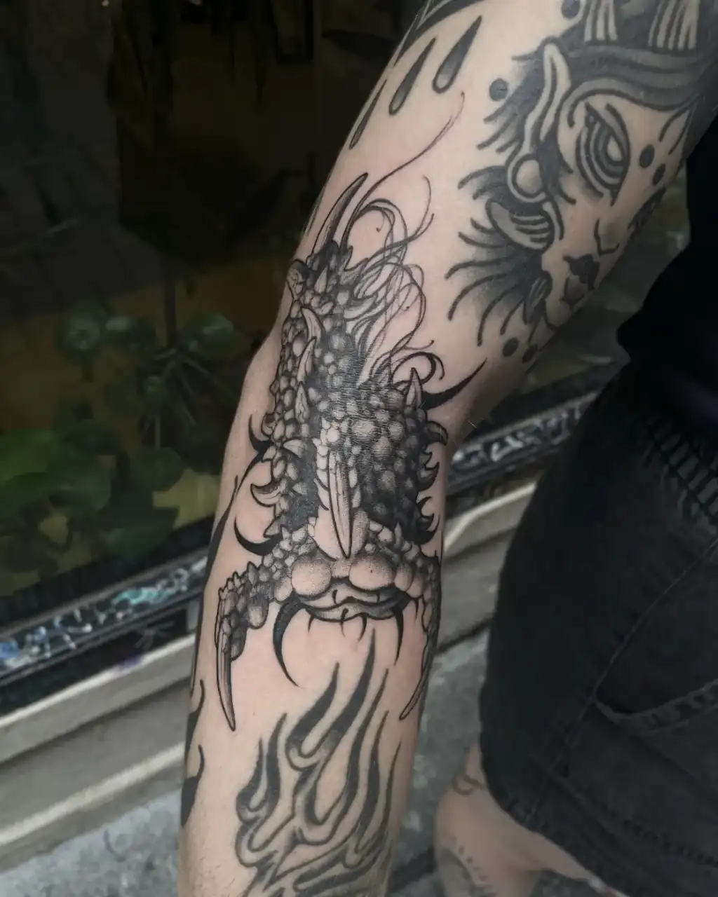 Codo y antebrazo recorridos por la cabeza acorazada de una criatura entre llamas, en black & grey con dotwork.