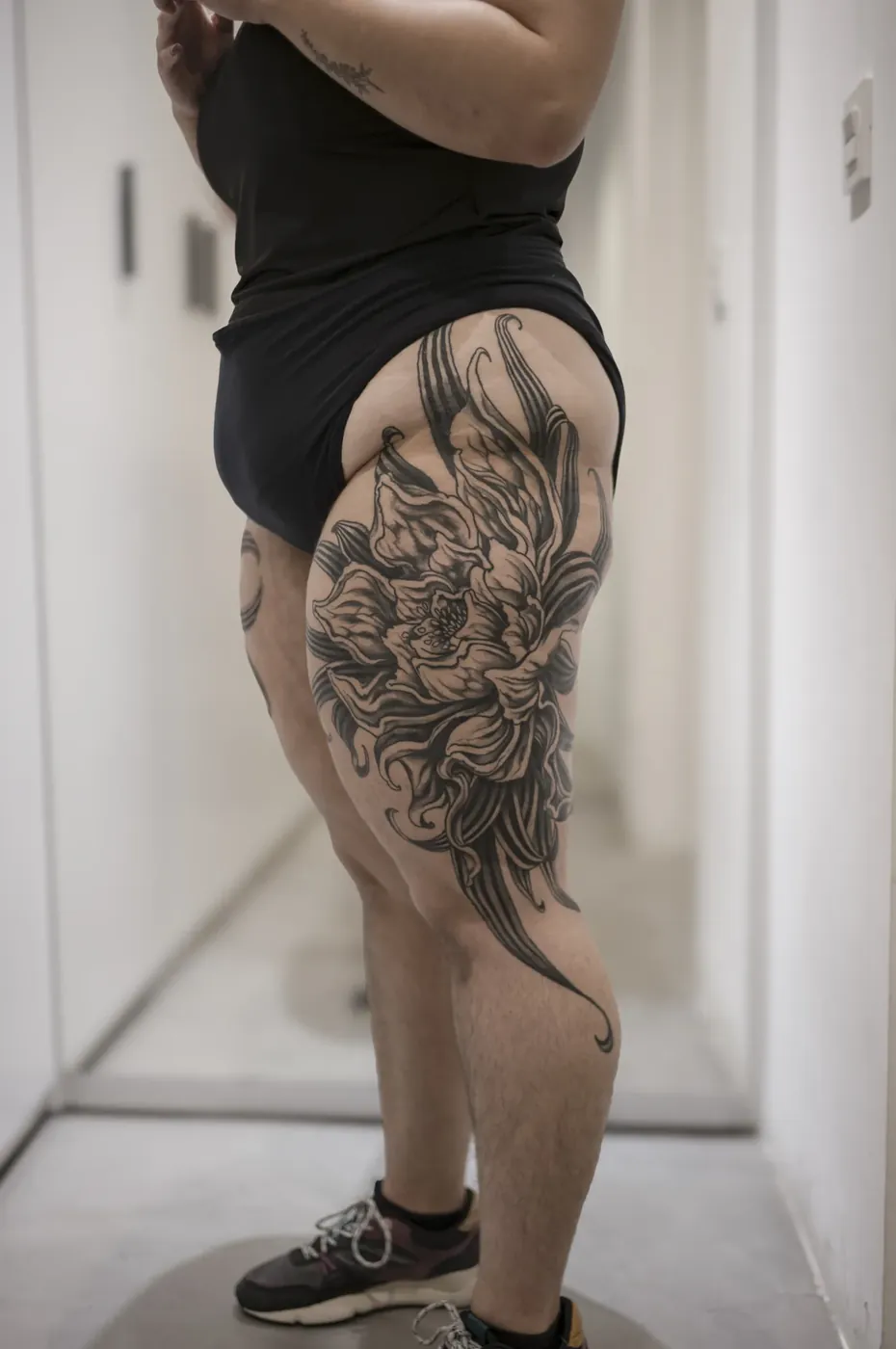 Cadera y muslo, de cuerpo entero: flor de pétalos largos en black & grey, con volutas que bajan hasta la rodilla.