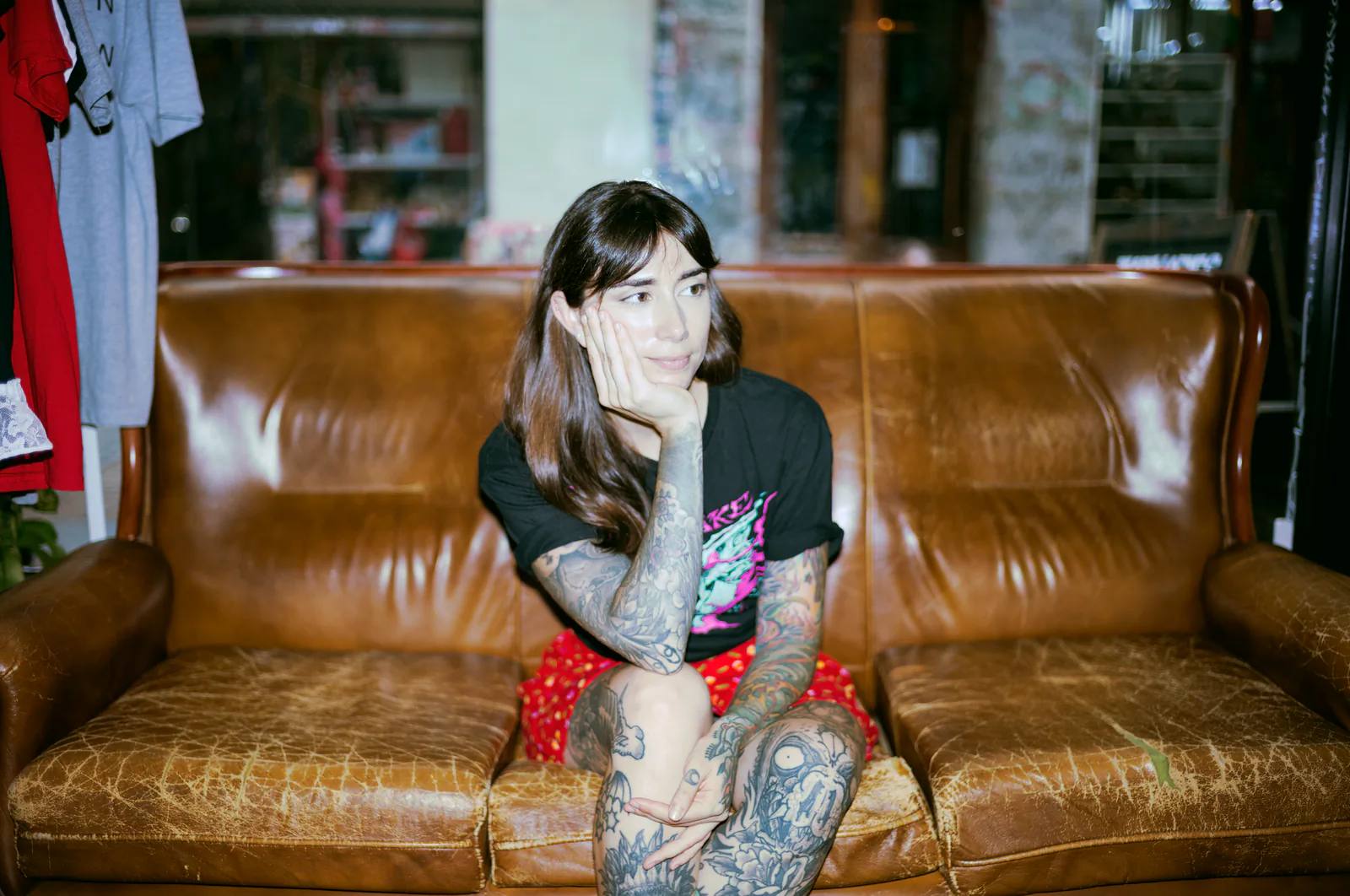 María sentada en un sofá de cuero marrón, brazos tatuados, mirando a cámara.