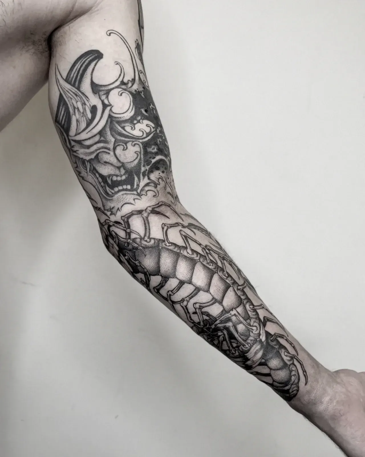 Manga completa en blackwork: una máscara hannya con olas ocupa el hombro y un ciempiés recorre el antebrazo hasta la muñeca. Fotografía en blanco y negro.