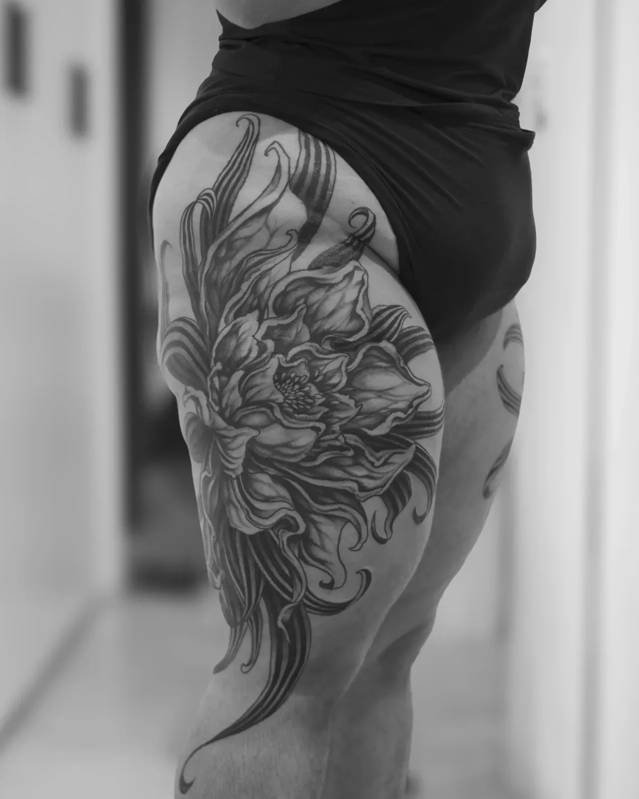 Crisantemo en blackwork sobre la cadera y el muslo, visto de perfil: la flor abre en el hueso de la cadera y las hojas bajan en tiras hasta la rodilla. Fotografía en blanco y negro.