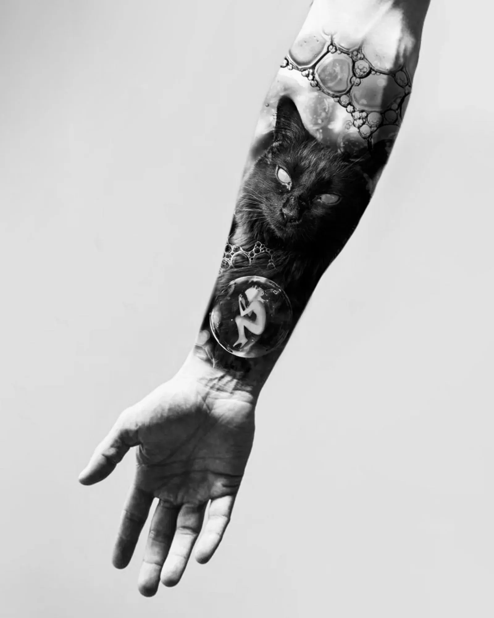 Diseño de realismo black & grey: un gato negro entre burbujas con una pequeña figura en una esfera, presentado en un montaje sobre un antebrazo.