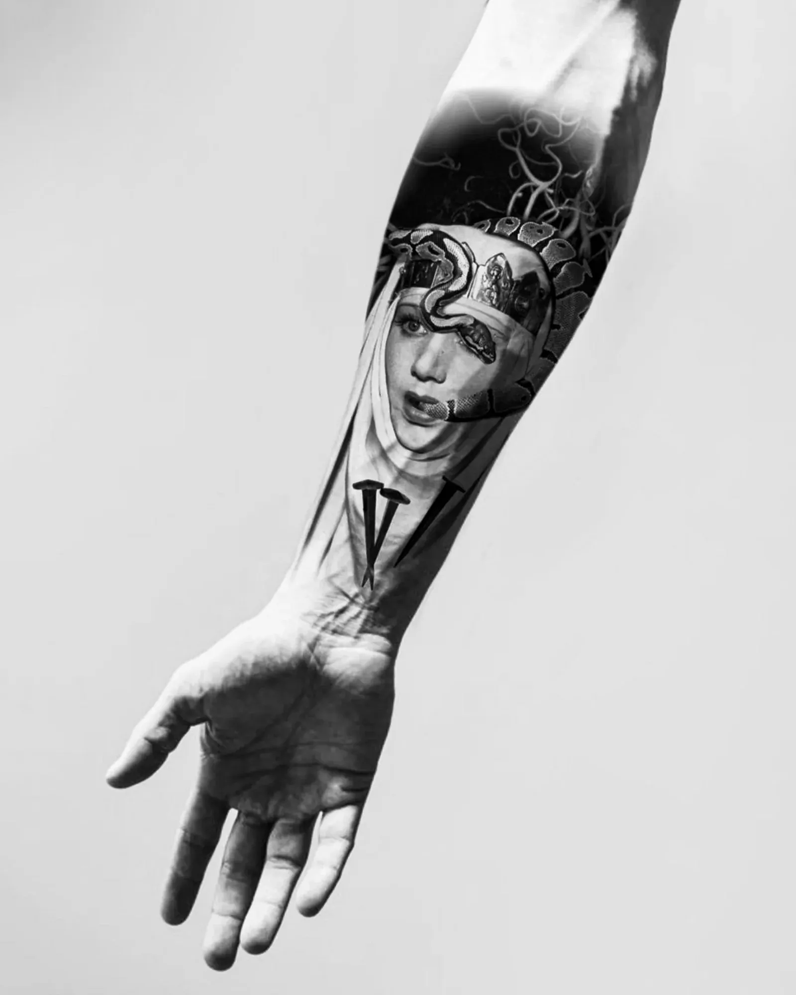 Diseño de realismo black & grey: un retrato femenino con toca, una serpiente sobre la cabeza y clavos cruzados, presentado en un montaje sobre un antebrazo.