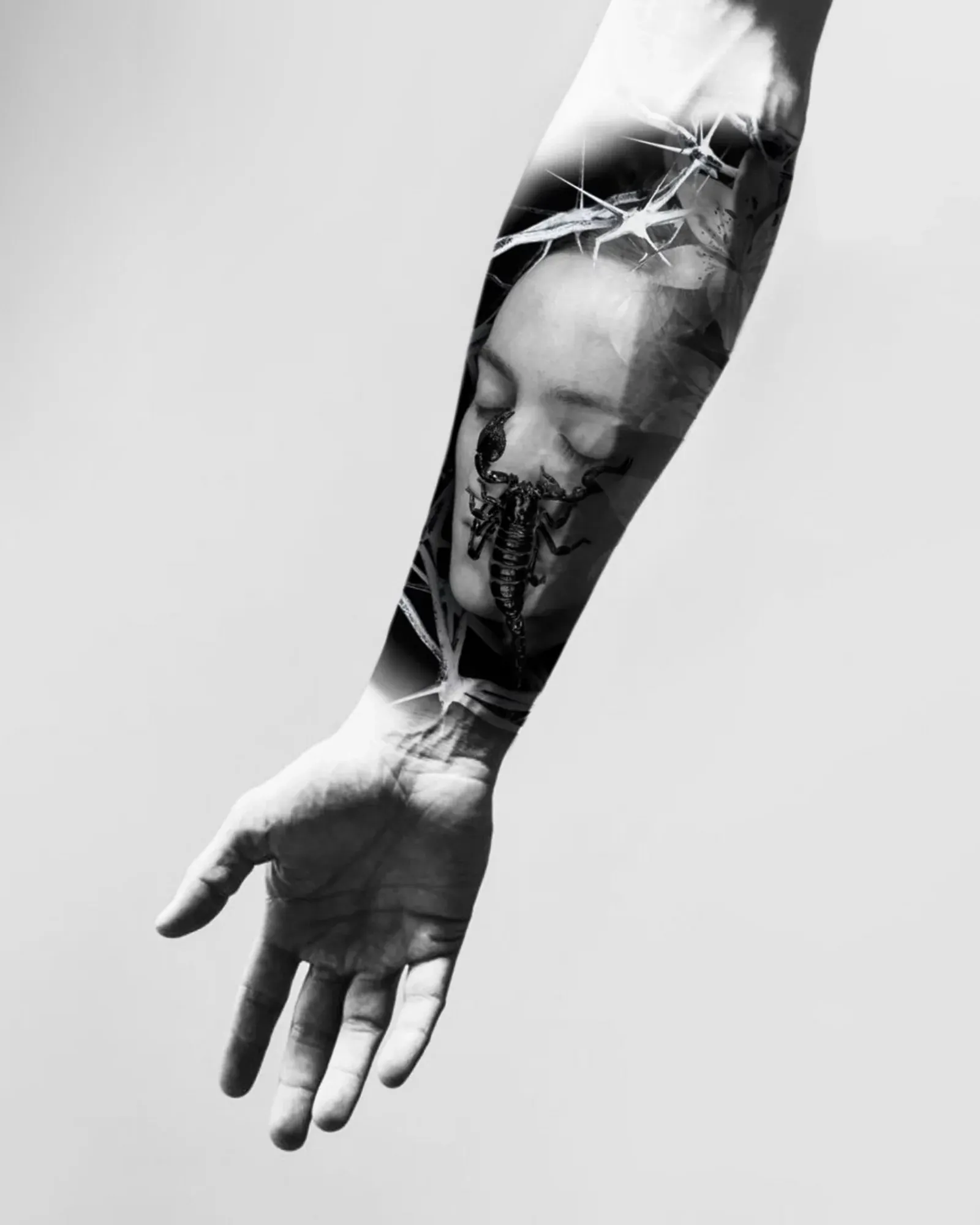 Diseño de realismo black & grey: un rostro de ojos cerrados con un escorpión y espinas, presentado en un montaje sobre un antebrazo.