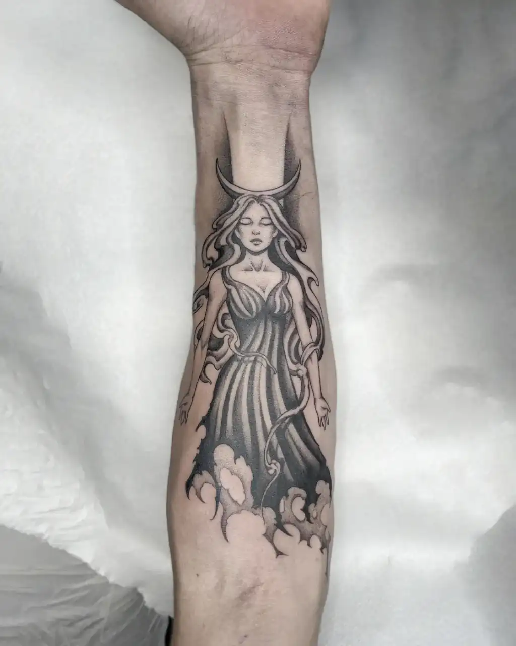 Antebrazo con una figura femenina de vestido largo bajo una luna creciente y sobre nubes, en black & grey de sombreado suave.