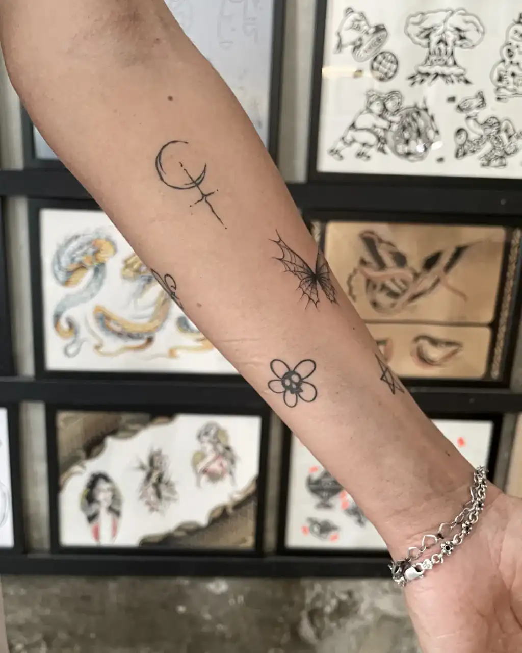 Antebrazo salpicado de tatuajes pequeños de línea fina: luna creciente, mariposa de telaraña y flor con calavera.