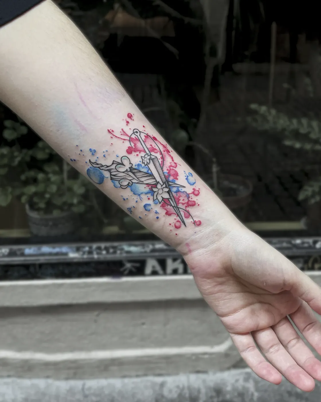 En el antebrazo, una daga rodeada de florecillas sobre salpicaduras rojas y azules, resuelta en acuarela.