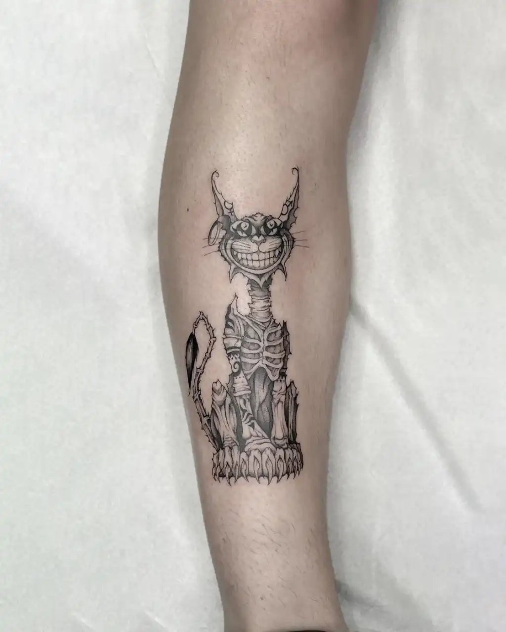 Antebrazo con un gato sonriente de cuerpo esquelético y remates ornamentales, en línea fina y sombras black & grey.