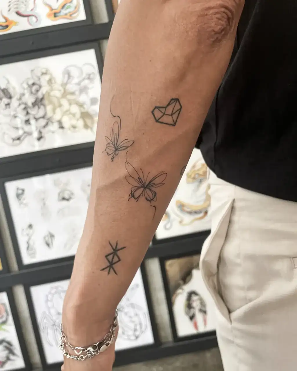 Sobre el antebrazo, varias piezas pequeñas de línea fina: dos mariposas, un corazón geométrico facetado y un símbolo de trazos rectos.