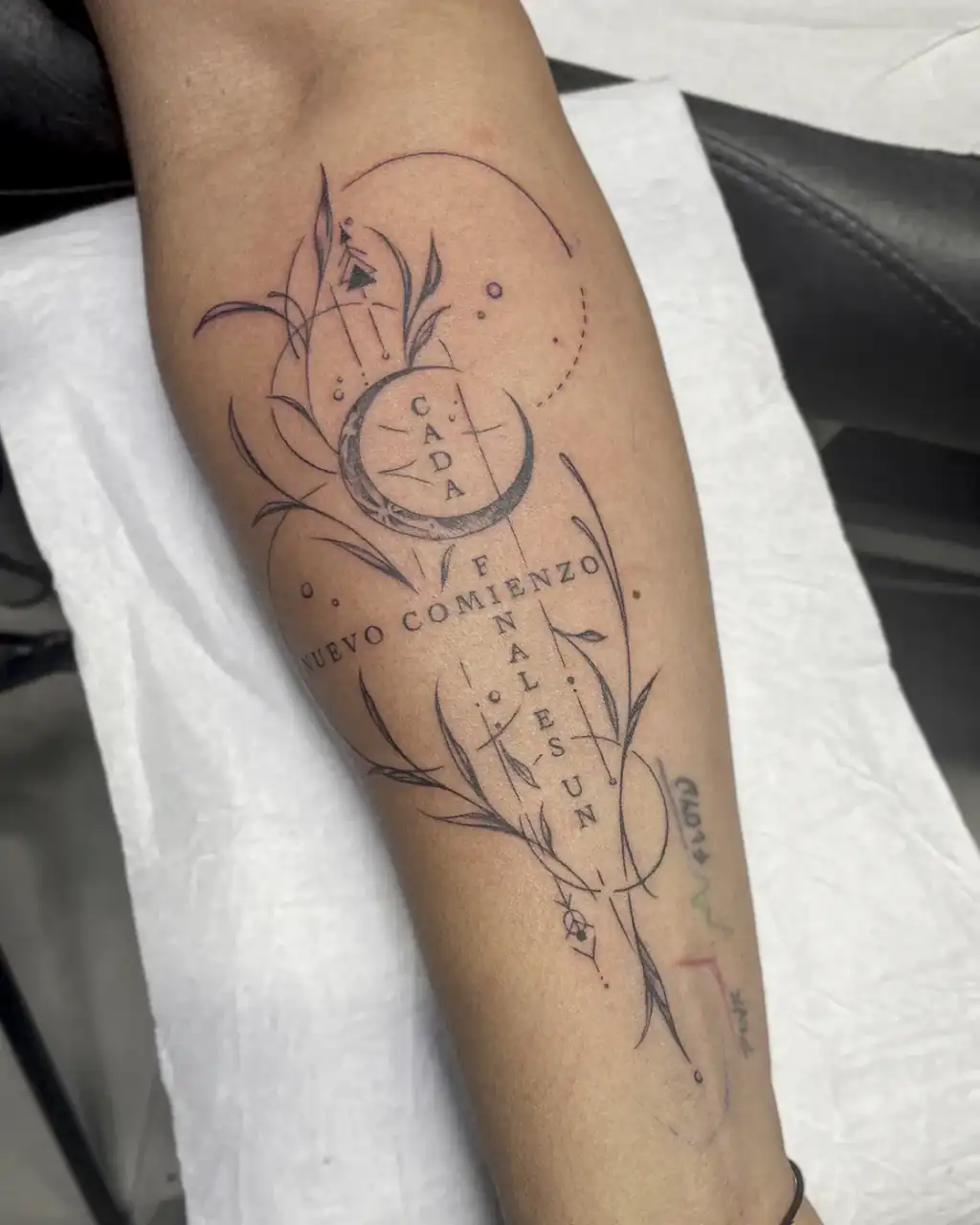 Antebrazo tatuado con luna, ramas y círculos de línea fina y el lettering «Cada final es un nuevo comienzo» en el centro.