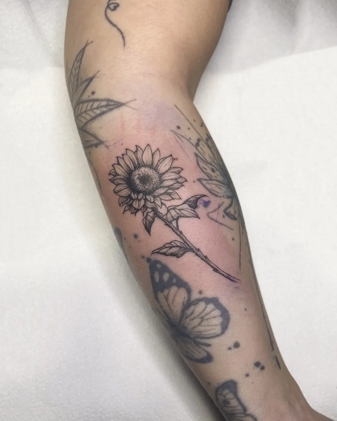 Brazo con un girasol de línea fina y centro en dotwork, entre mariposas y hojas ya tatuadas.