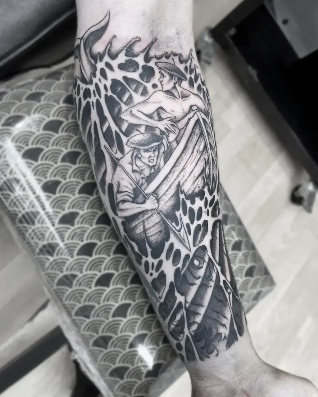 Antebrazo donde dos figuras de sombrero asoman entre escamas y formas orgánicas, resueltas en blackwork y grises.