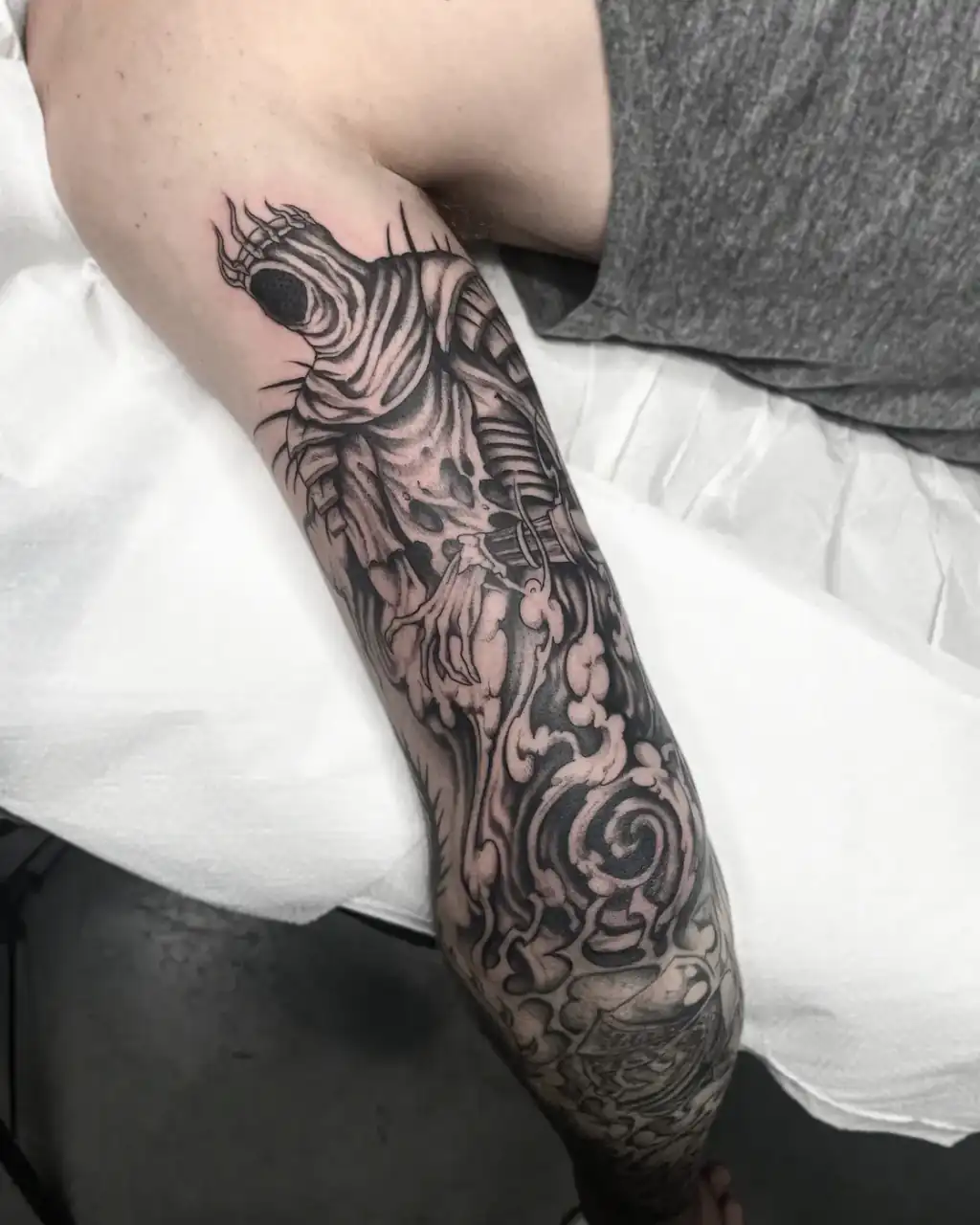 Del hombro a la mano, una figura encapuchada de mano esquelética entre nubes y volutas de black & grey.