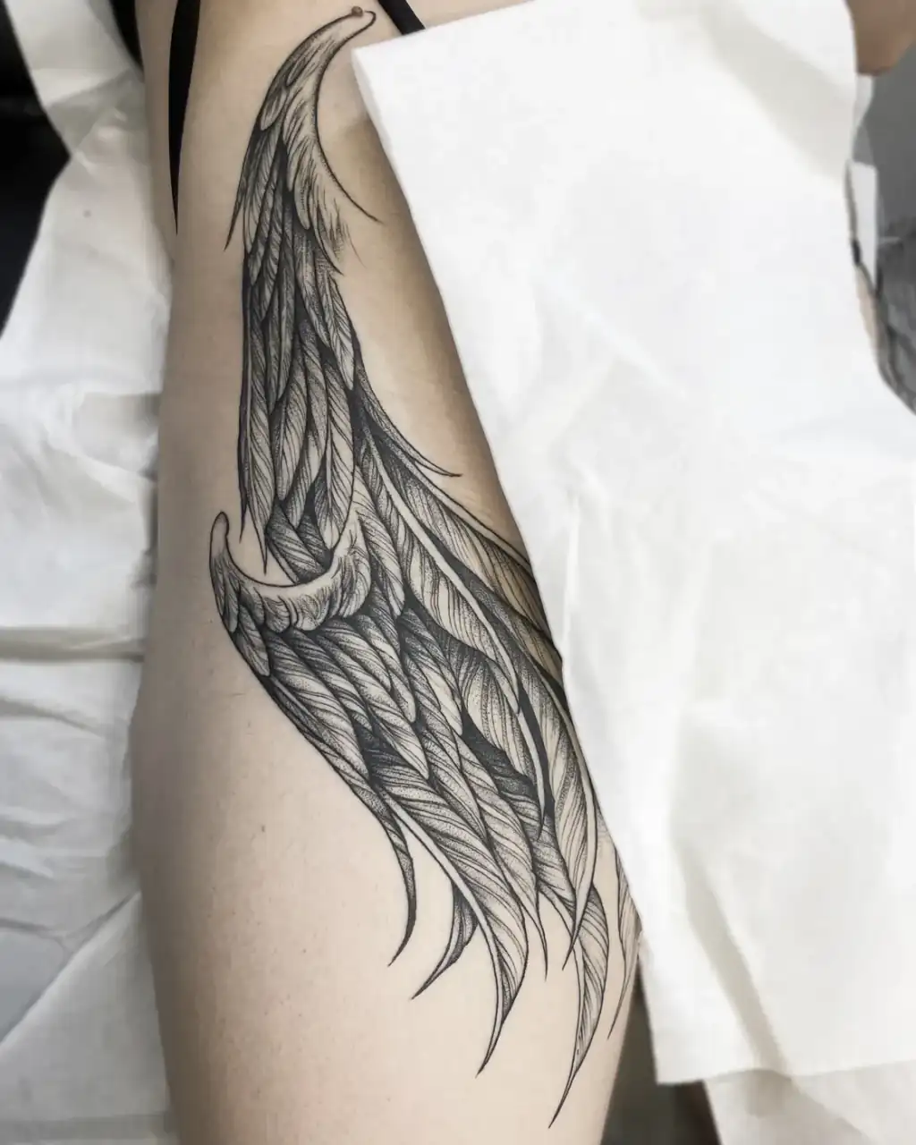 Muslo con un ala de plumas de gran tamaño, dibujada a línea fina y sombreada en dotwork.