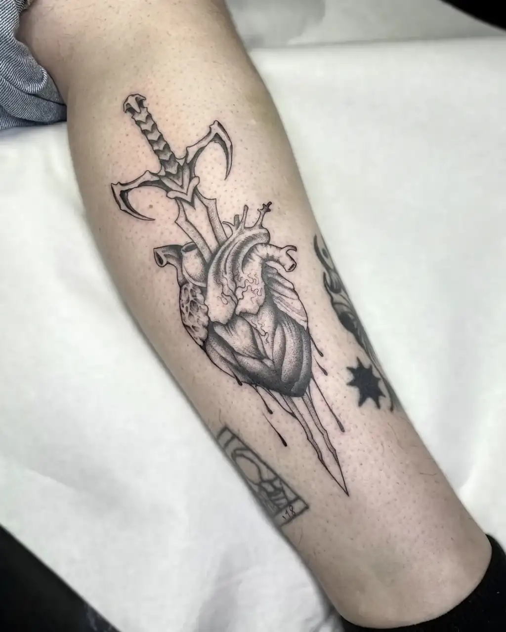 En la pierna, un corazón anatómico atravesado por una daga gotea tinta; black & grey con textura de dotwork.