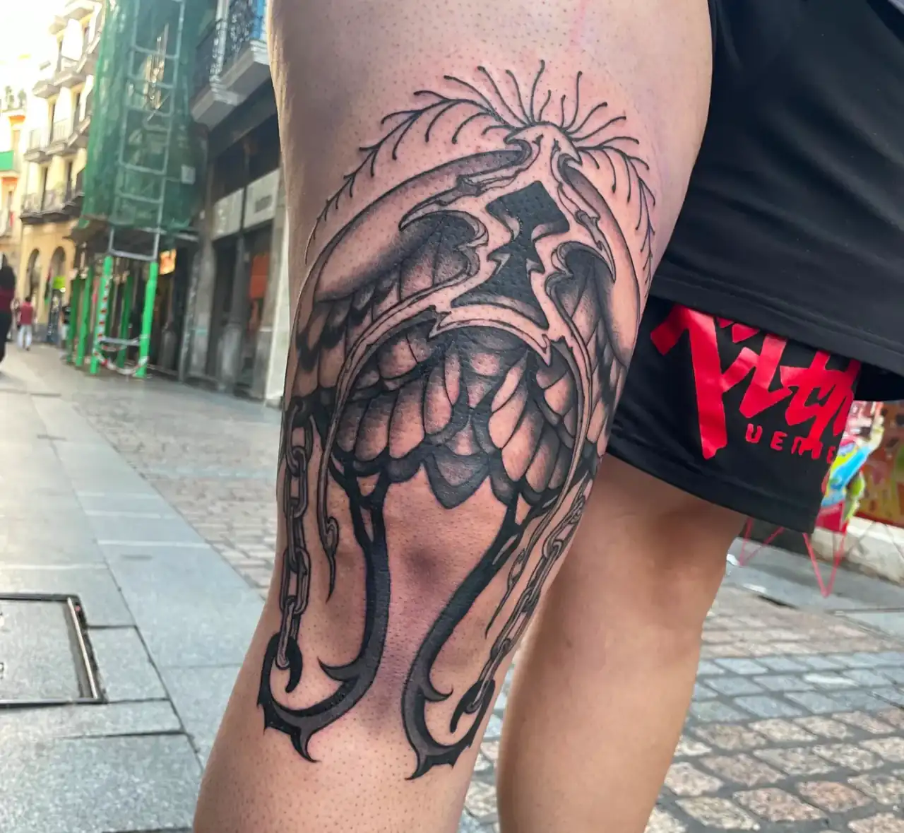 Rodilla envuelta por una criatura alada con escamas, cadenas y púas, en blackwork macizo con remates neotribales.